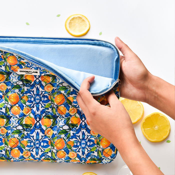 Hands opening a yellow lemon-patterned quilted laptop sleeve with blue lining; lemon slices on a white surface nearby.