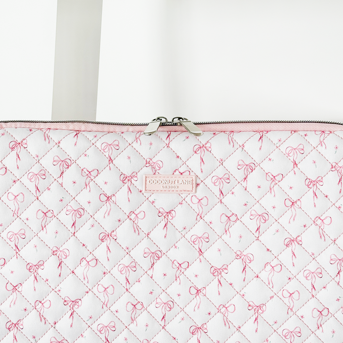 Pink quilted laptop sleeve with dainty bow pattern, diamond stitching, zipper closure, and a centered label.