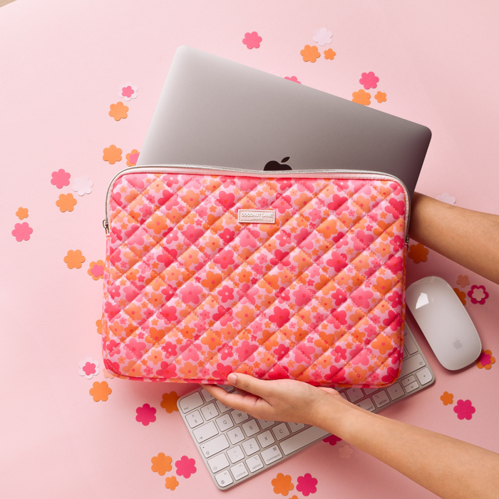 Hands hold a quilted pink and orange floral laptop sleeve above a MacBook, mouse, and keyboard on a pink surface.