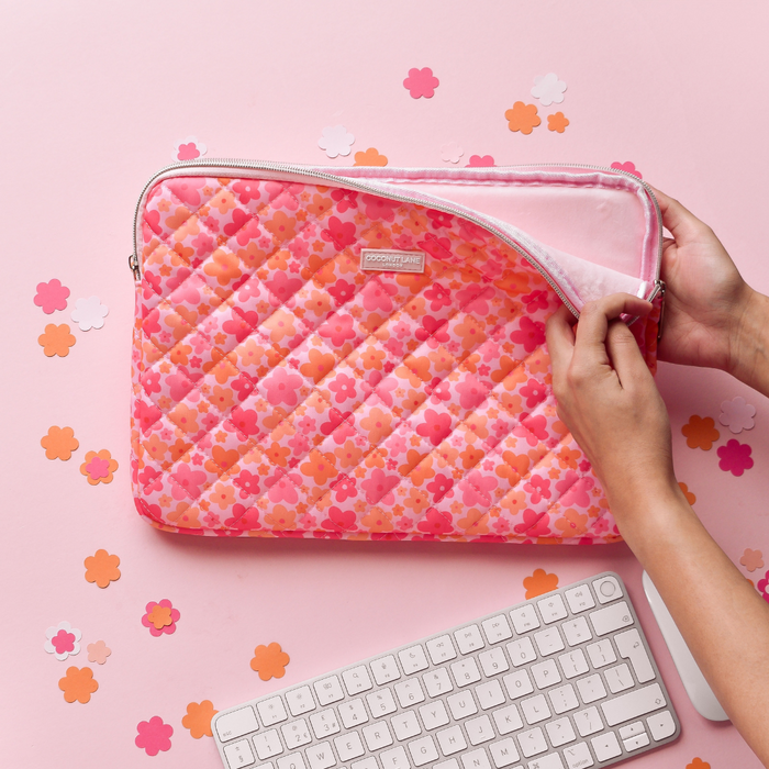 Hands opening a pink and orange quilted laptop sleeve on a pink desk with confetti and a keyboard nearby.