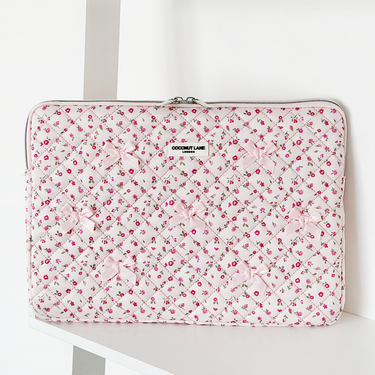 Pink quilted laptop sleeve with watercolor flower pattern, bows, and zipper, displayed on a white shelf.