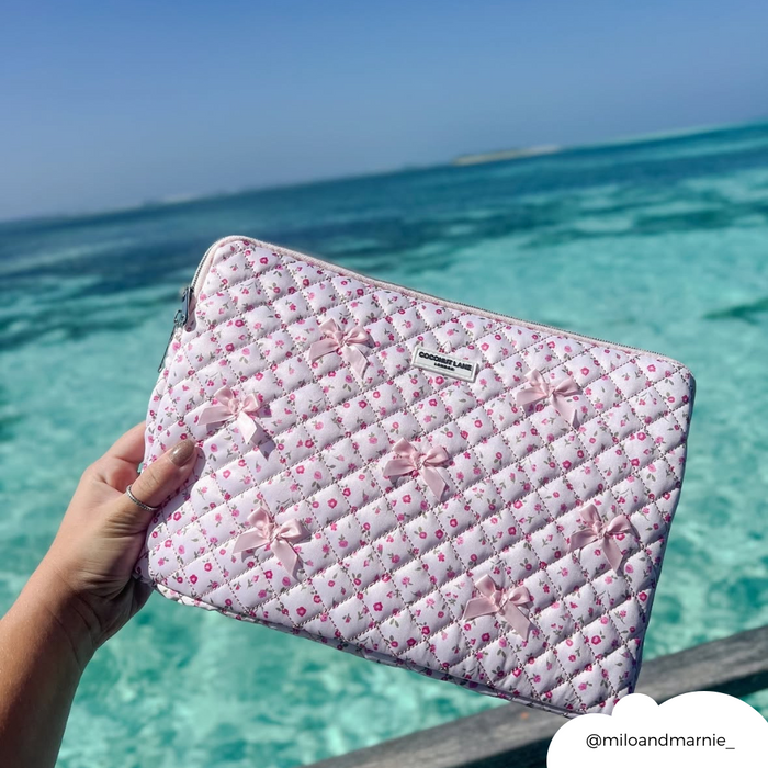 Hand holding a pink quilted laptop sleeve with a watercolour flower pattern near turquoise ocean and clear sky.