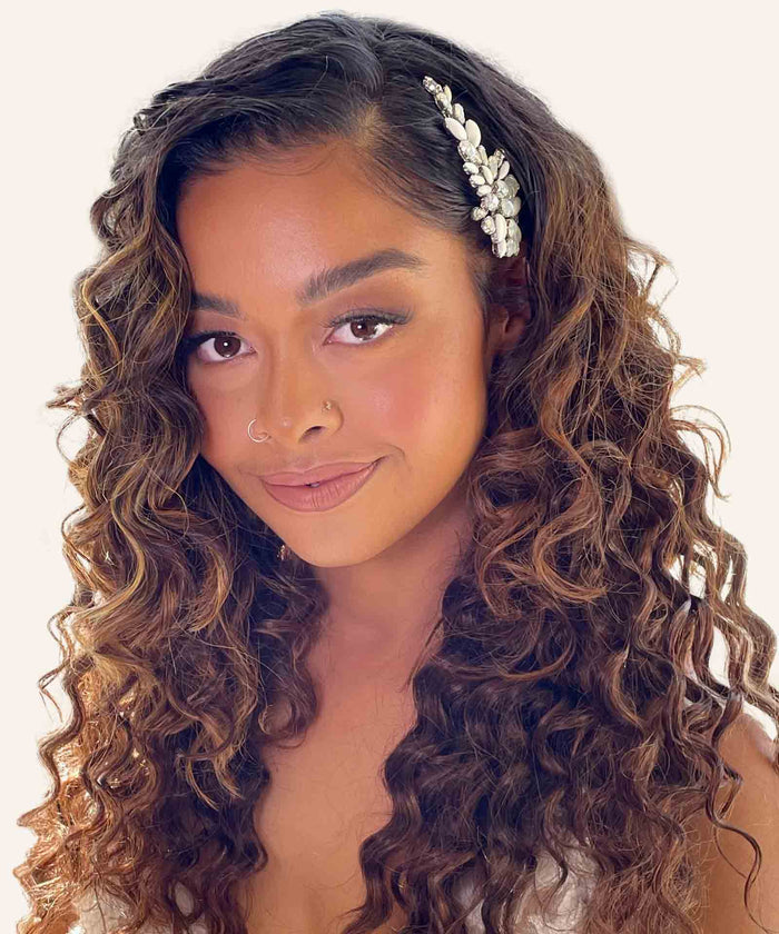 A woman with long curly brown hair wears a pearl and crystal hair accessory, nose ring, subtle makeup, and smiles gently.