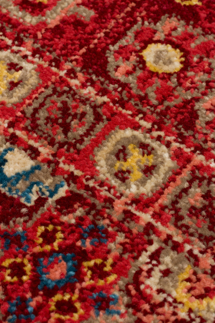 Close-up of a deep red rug with a repeating geometric medallion pattern in beige, yellow, and blue.