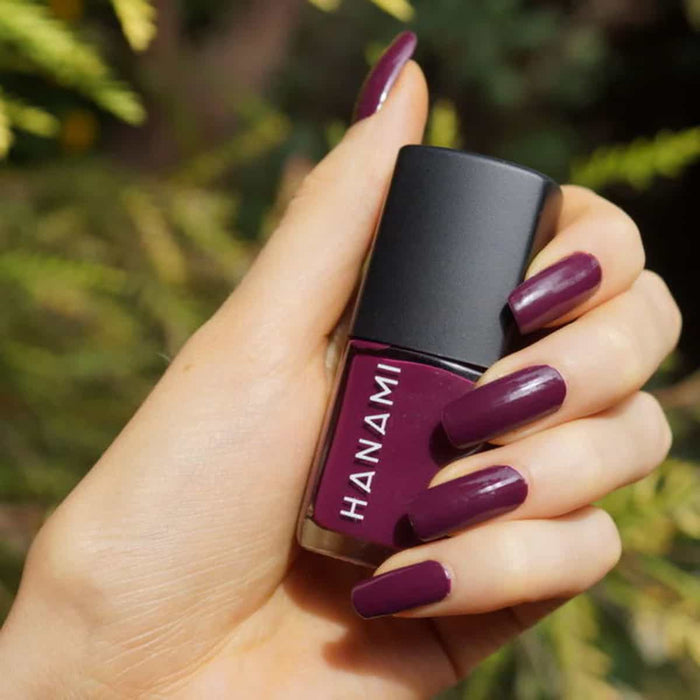 Hand holding a bottle of Hanami nail polish with matching purple nails against a blurred green background.
