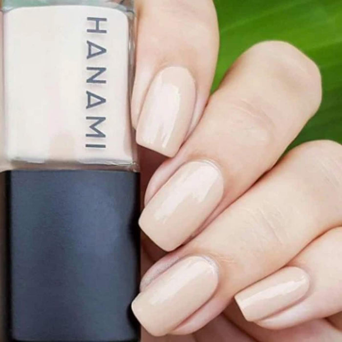 Nail polish bottle with 'HANAMI' branding next to a hand with beige nail polish on a green leaf background.