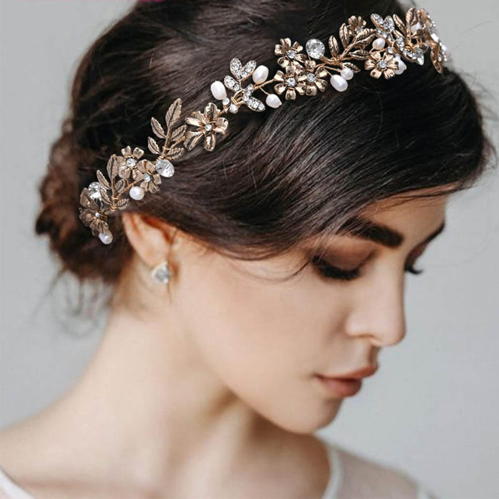 A woman with dark hair in a low bun wears a gold floral headband with pearls and crystals, pearl drop earrings, and gazes down softly against a softly blurred, neutral background.