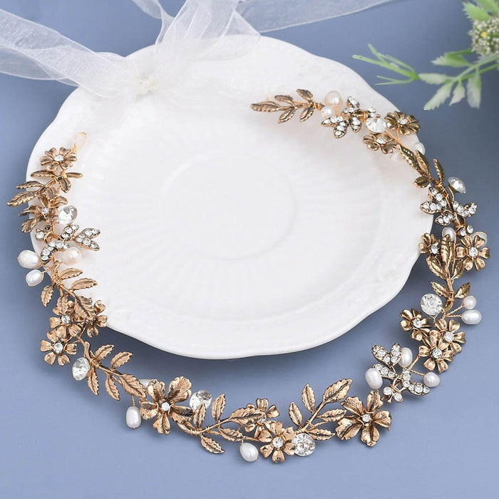 A delicate gold floral headpiece with crystal gems and pearls rests on a white plate, sheer ribbon, and green sprig on light blue.