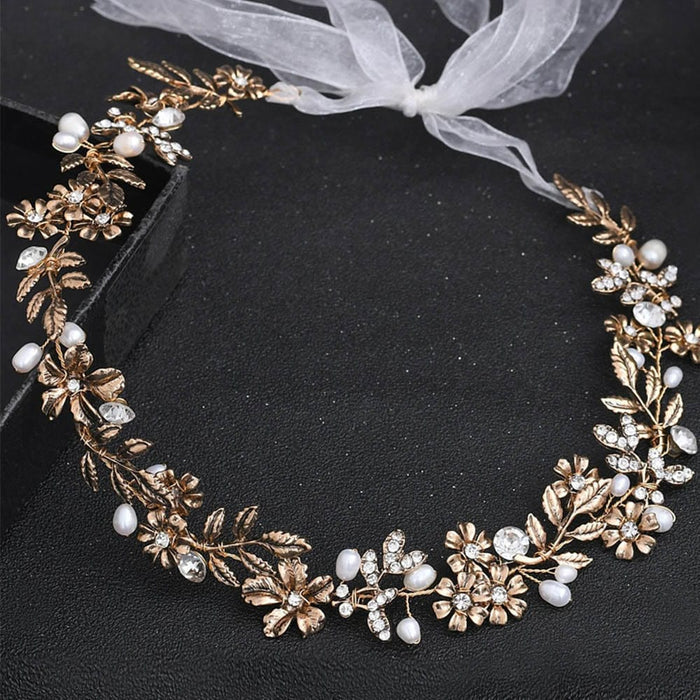 A delicate gold floral headpiece adorned with white pearls, clear crystals, and intricate leaf designs tied with a sheer white ribbon on a black textured surface.