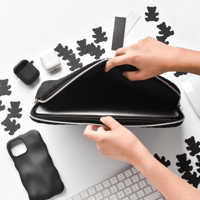 Hands setting down a beige teddy-textured laptop sleeve on a white desk with bear cutouts and phone cases nearby.