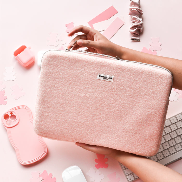 Hands holding a beige Teddy Love-a-Lot laptop sleeve on a pink desk with keyboard, phone case, and scrunchie nearby.