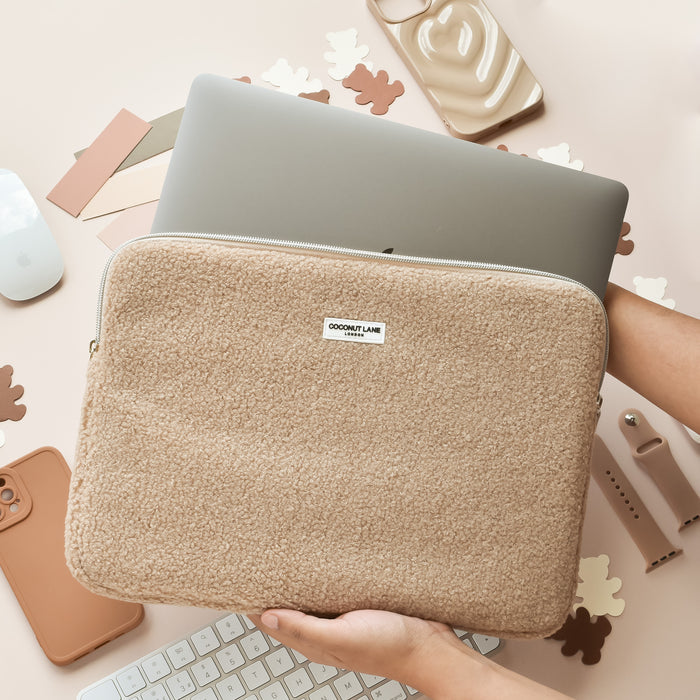 A hand slides a laptop into a tan, fuzzy Coconut Lane Teddy Rupert laptop sleeve on a desk with tech items and color swatches.