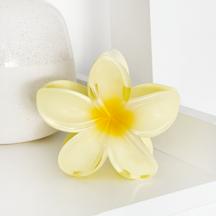 Butter yellow flower-shaped claw clip on a white shelf beside a white ceramic vase.