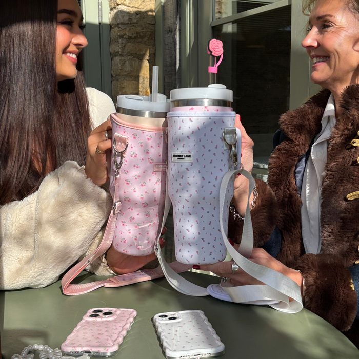 Two women with Coconut Lane white floral tumbler carry cases; phones on a sunlit table.