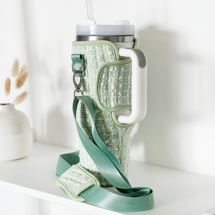 Matcha green textured tweed tumbler carry case with strap and handle on a white shelf beside a vase.