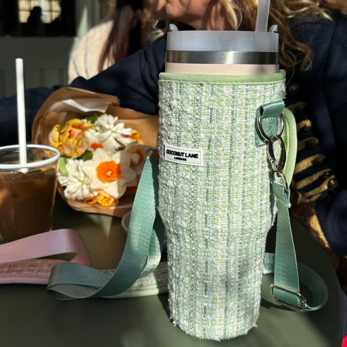Green textured tweed tumbler carry case with a Coconut Lane travel mug on a sunny table next to flowers and iced coffee.