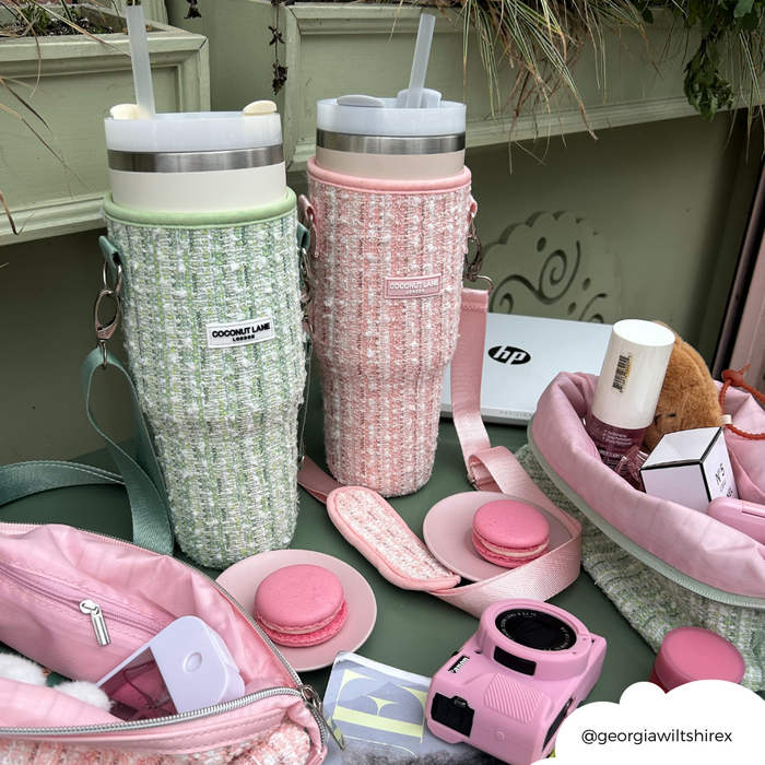 Two pastel tumblers in green textured tweed carry cases, on a table with pink bags and assorted macarons.