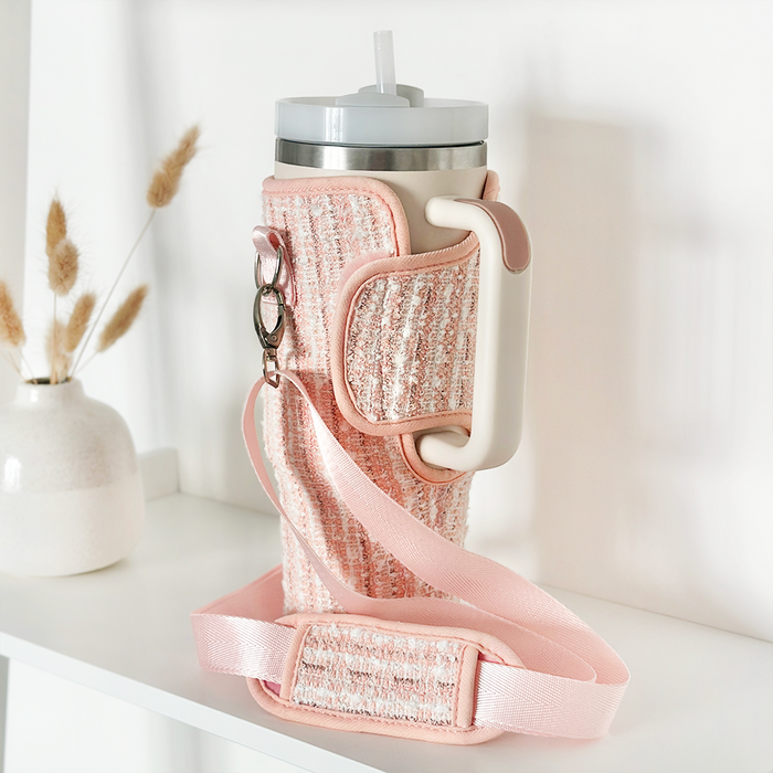 Pink textured tweed tumbler carry case with handle and strap on a white shelf next to dried flowers.