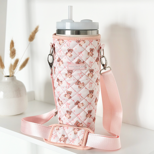 Quilted beige Teddy tumbler carry case with strap on a white shelf next to a vase of dried grass.