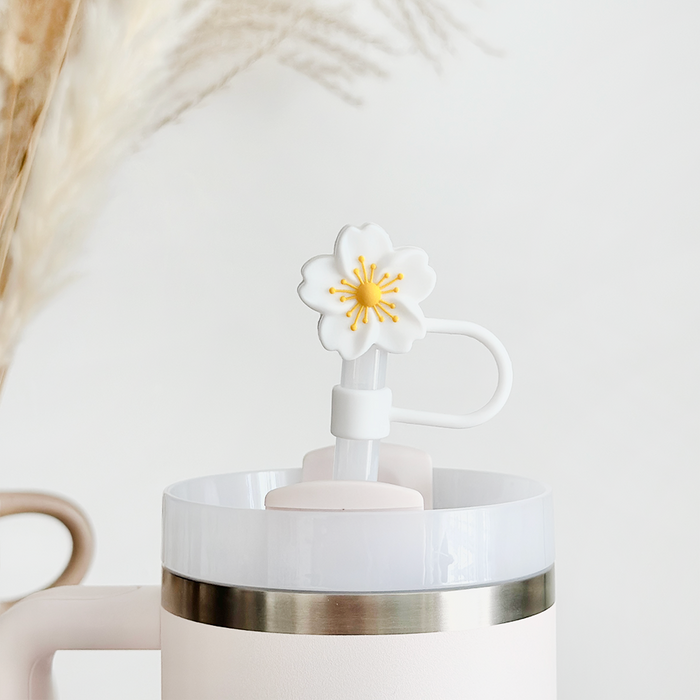 White tropical flower straw cover on a white cup with steel rim, placed on dried grass with a neutral background.