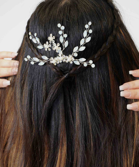A person with long brown hair in half-up braids, wearing a hair accessory with pearl-like beads and silver leaves, adjusts their boho hairstyle.