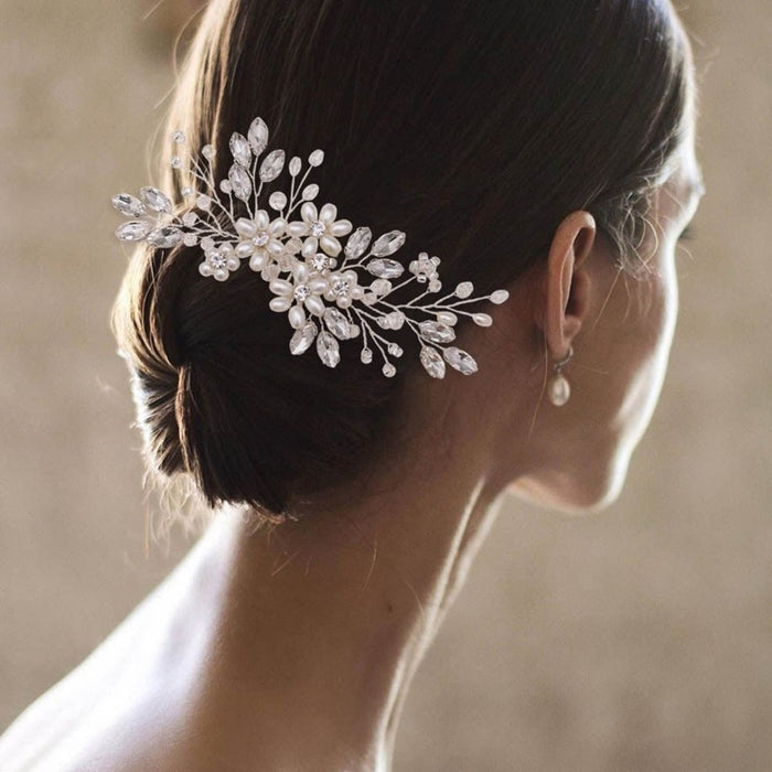 A woman with dark hair in a low bun wears a pearl and crystal hairpiece and pearl drop earring. Blurred background enhances elegance.