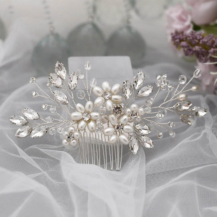 A decorative bridal hair comb with pearls and crystal beads in a floral design on soft white tulle, blurred flowers and glass behind.